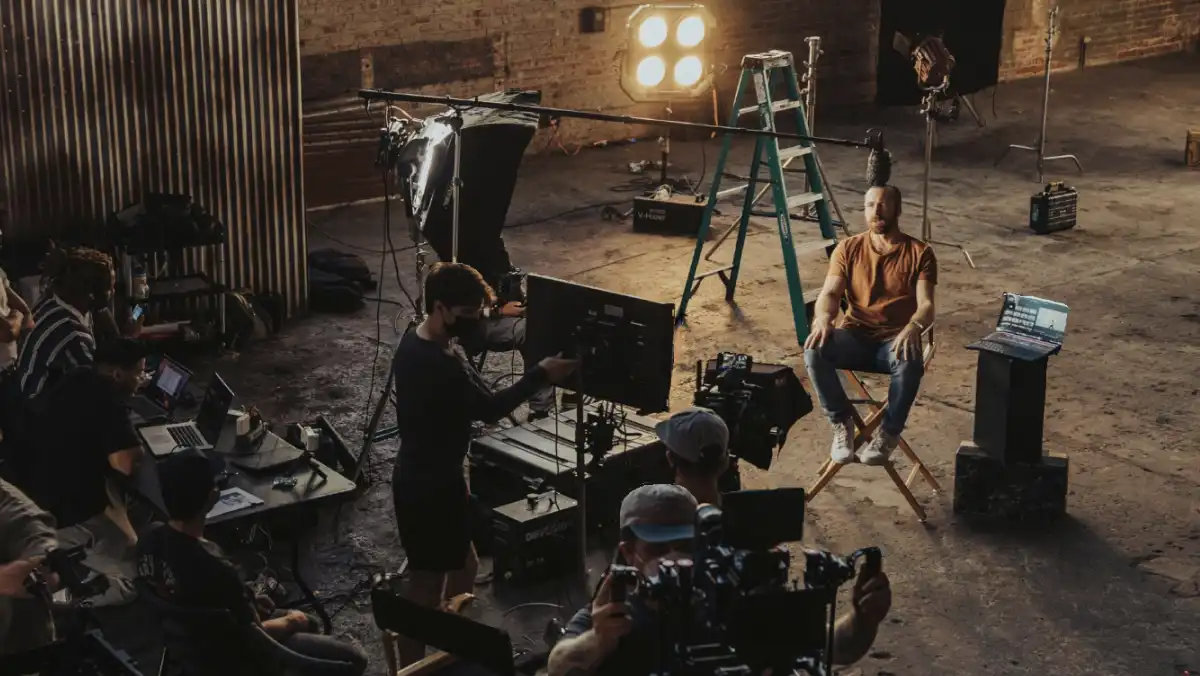 Behind-the-scenes view of a video production set, with a crew filming a man seated on a director's chair under professional lighting. Cameras, monitors, laptops, and lighting equipment fill the industrial-style space. This image is part of a template designed to showcase media and creative industry events.